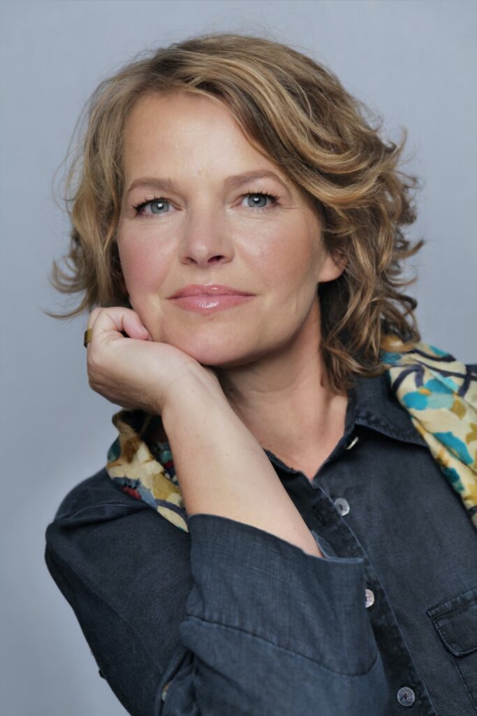 Portrait of a woman with blond, wavy hair resting her chin on her hand, wearing a dark denim shirt and colorful scarf.
